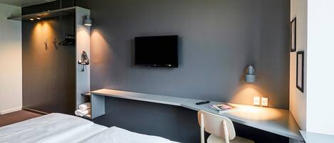 A modern hotel room with a bed, a desk with a chair, a flat-screen TV, and a wall-mounted shelf.