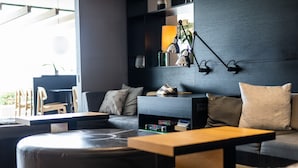 Una sala moderna con un sofá, una otomana y una mesa de centro. Se observa una estantería con libros y objetos decorativos. Hay una lámpara fijada a la pared, y una planta se ve al fondo.