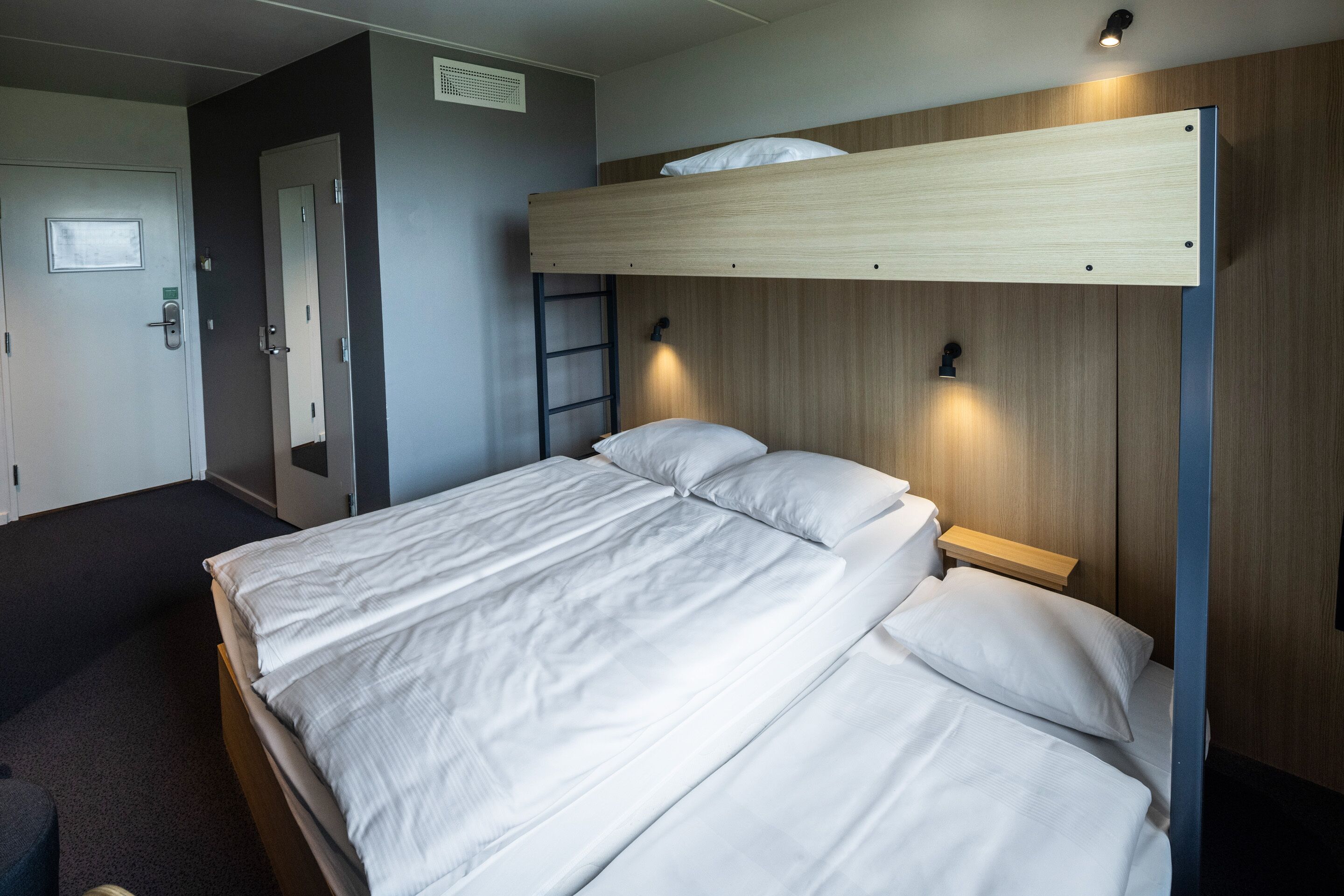 A bunk bed with a wooden headboard and a small bedside table. There is a door with a mirror and a light switch on the wall.