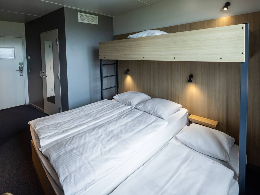 A bunk bed with a wooden headboard and a small bedside table. There is a door with a mirror and a light switch on the wall.