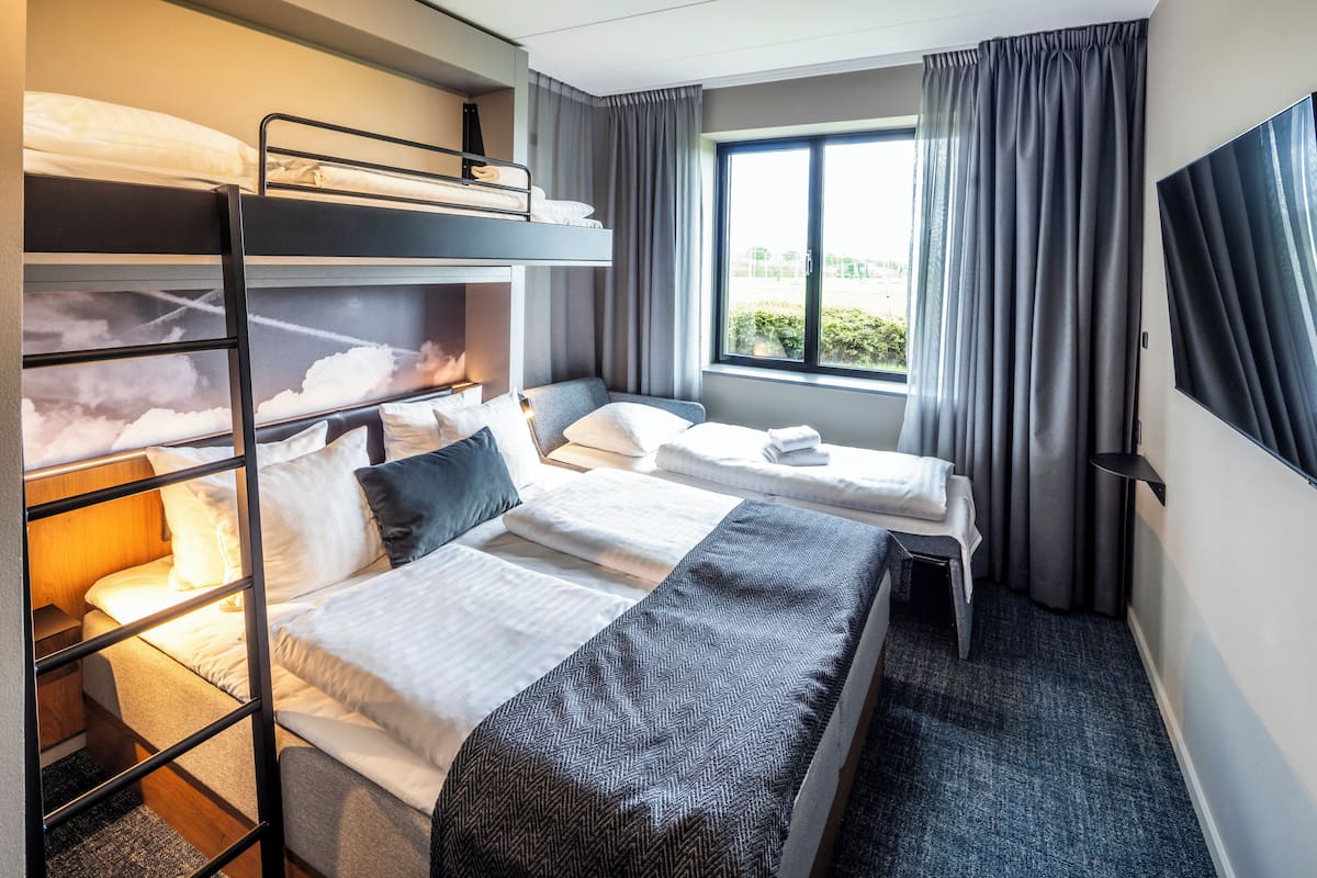 A hotel room with bunk beds, a desk, a chair, a window with curtains, and a flat-screen TV.