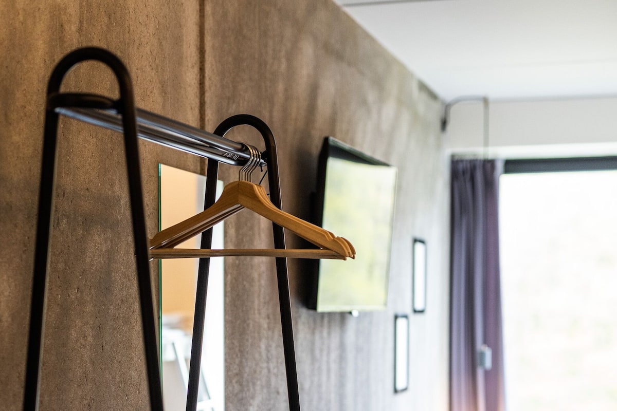 A modern hotel room with a concrete wall, a flat-screen TV mounted above a bed, a wooden hanger on a black metal stand, a round ottoman, and a window with curtains.