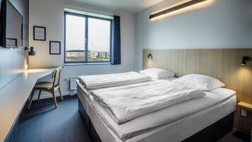 A hotel room with a bed, a desk, and a chair. There is a window with a view of buildings outside.