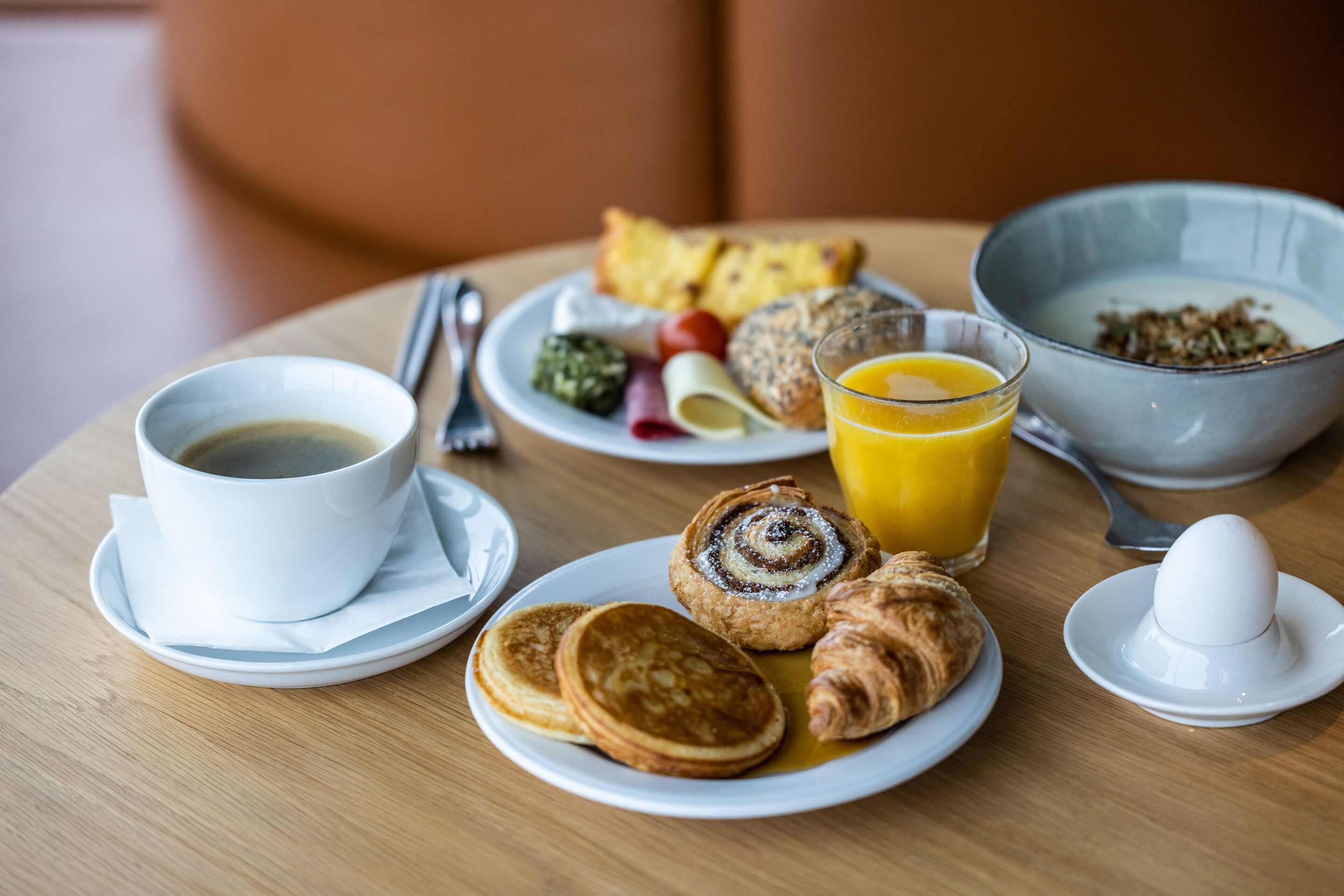 A breakfast table with pancakes, croissants, a bowl of cereal, a glass of orange juice, and a cup of coffee.