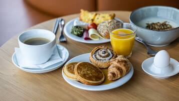 A breakfast table with pancakes, croissants, a bowl of cereal, a glass of orange juice, and a cup of coffee.