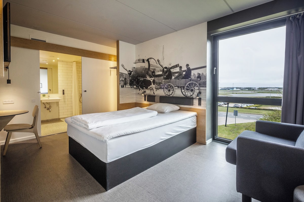 A hotel room with a bed, a desk, a chair, a large window with a view of an airport, and a mural on the wall.