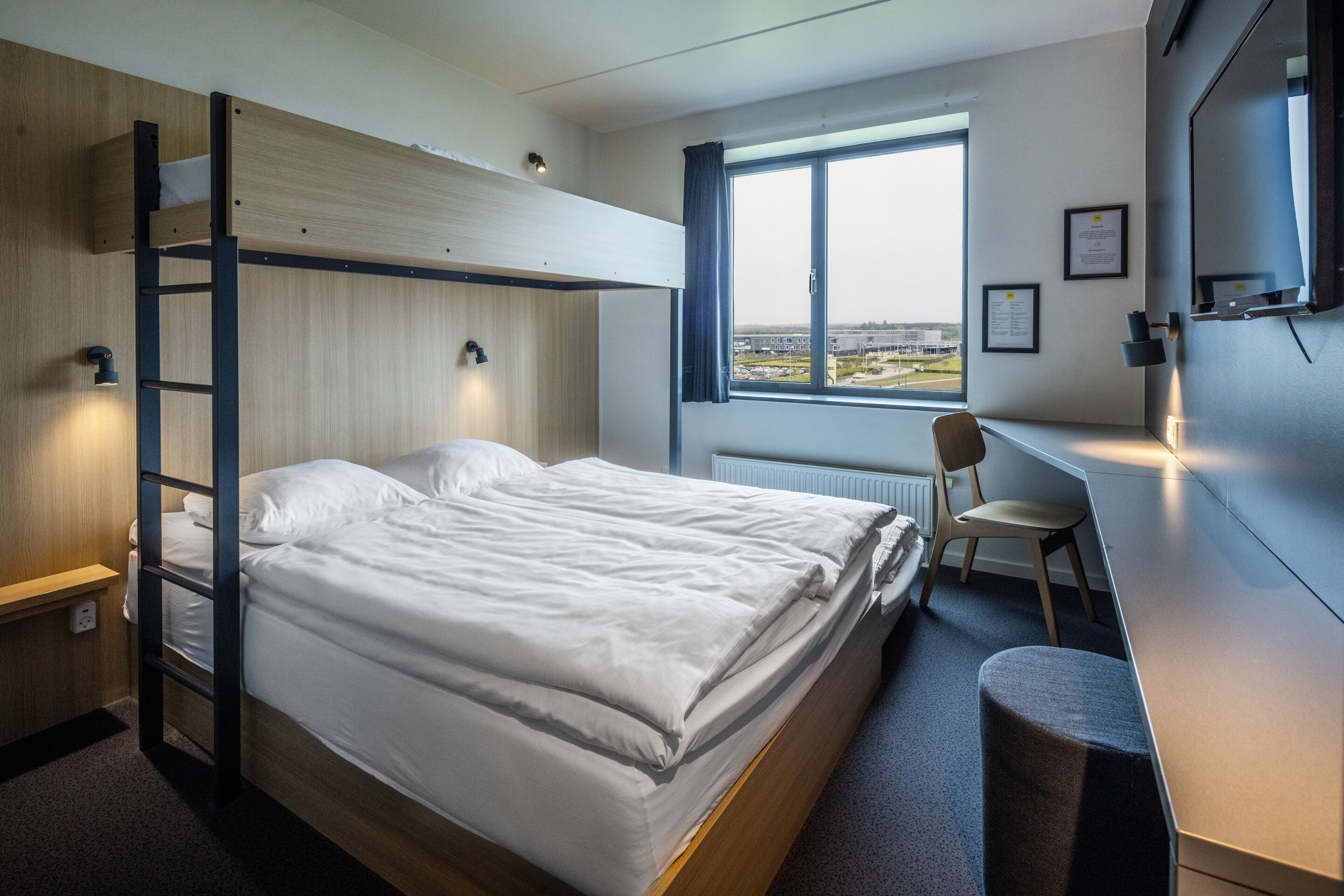 A bunk bed with a desk and chair, a window with a view, and a wall-mounted TV.