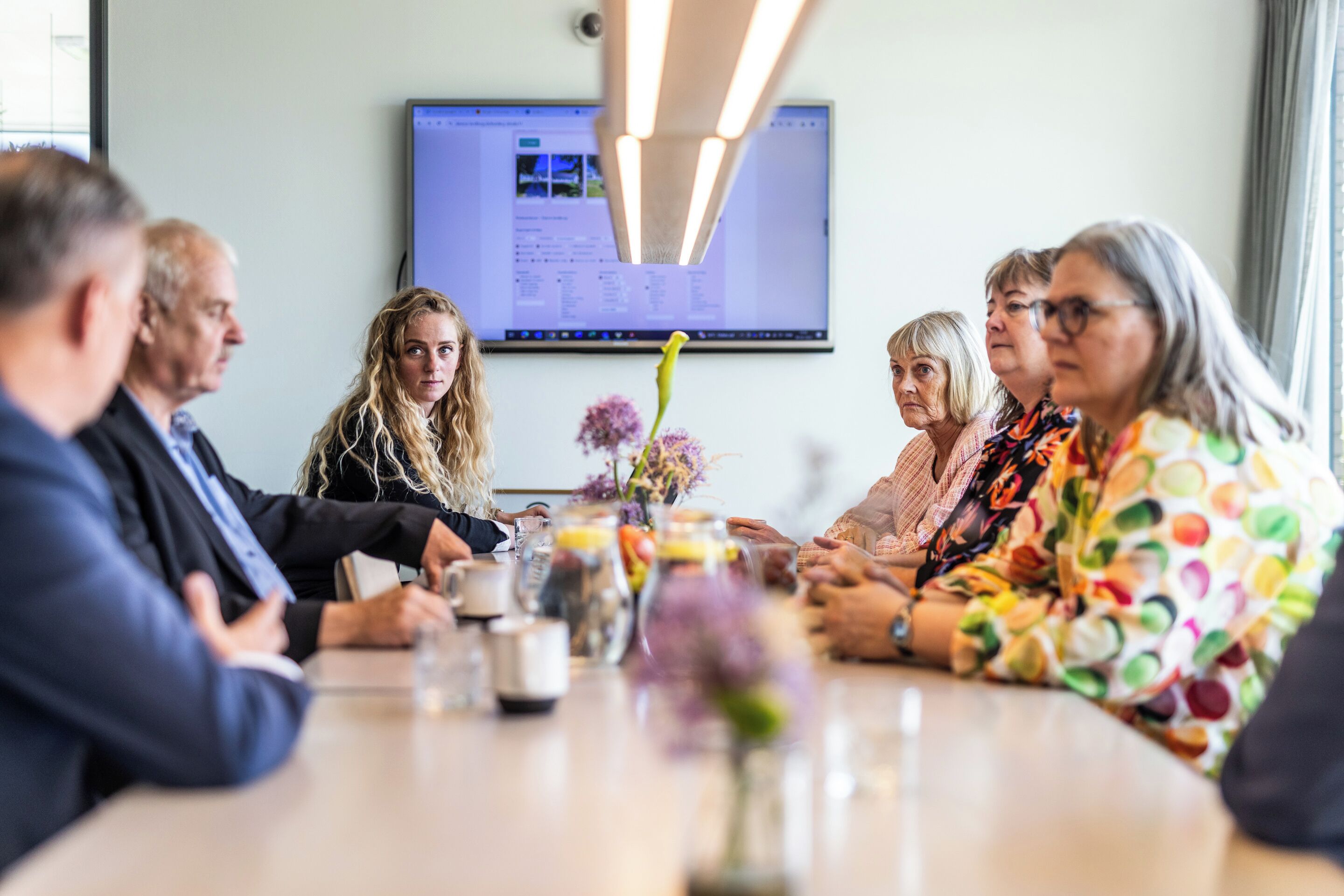 A meeting room with a large screen displaying a presentation, a table with a vase of flowers, and attendees seated around it.