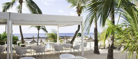 A beachside outdoor seating area with white tables and chairs, palm trees, and a view of the ocean.