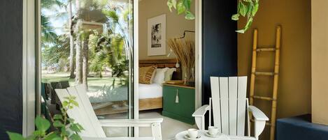 A room with a bed, a green cabinet, a white chair, a table with cups, and a view of palm trees through a glass door.