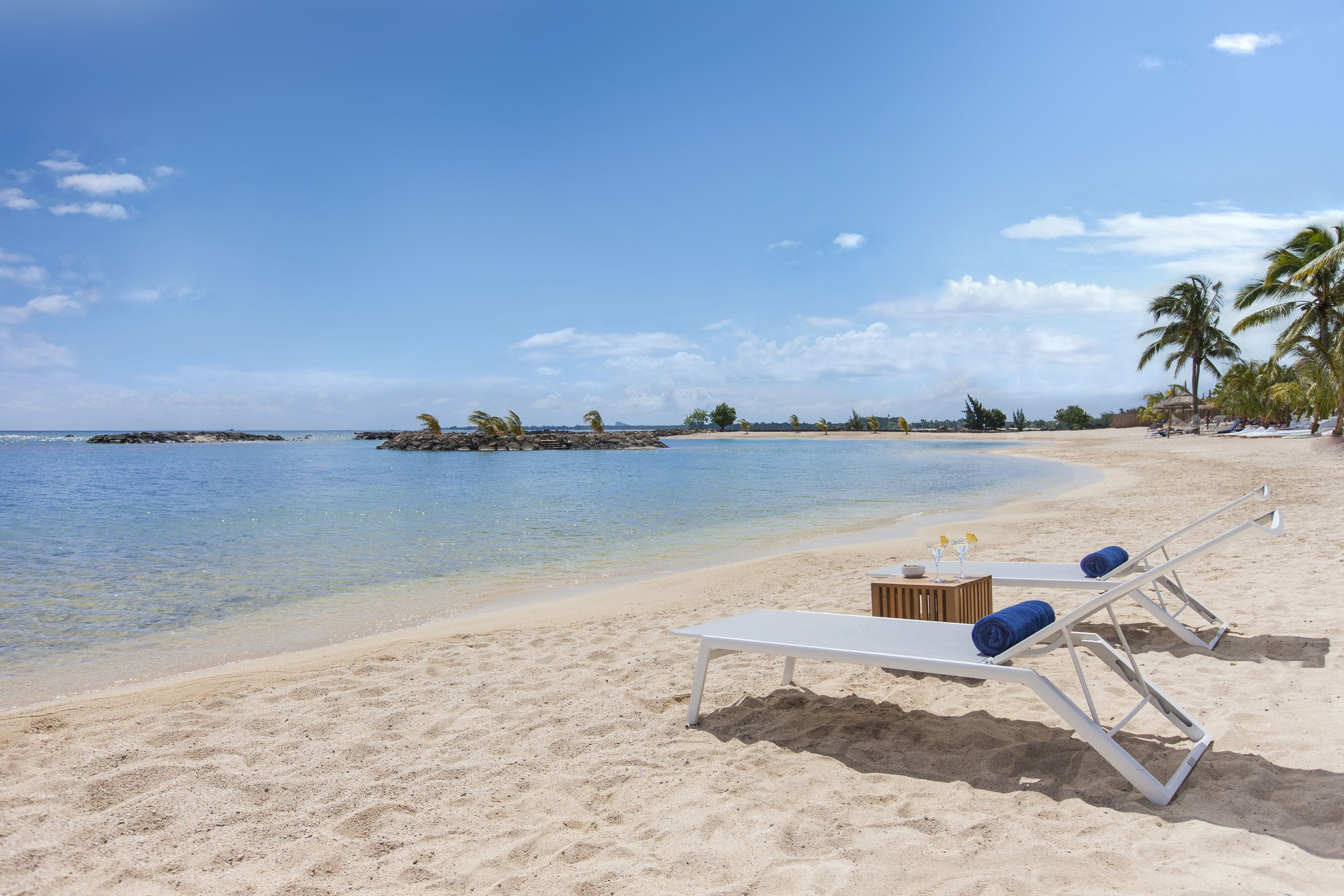 A beach with white lounge chairs and blue cushions, a wooden table, and palm trees.