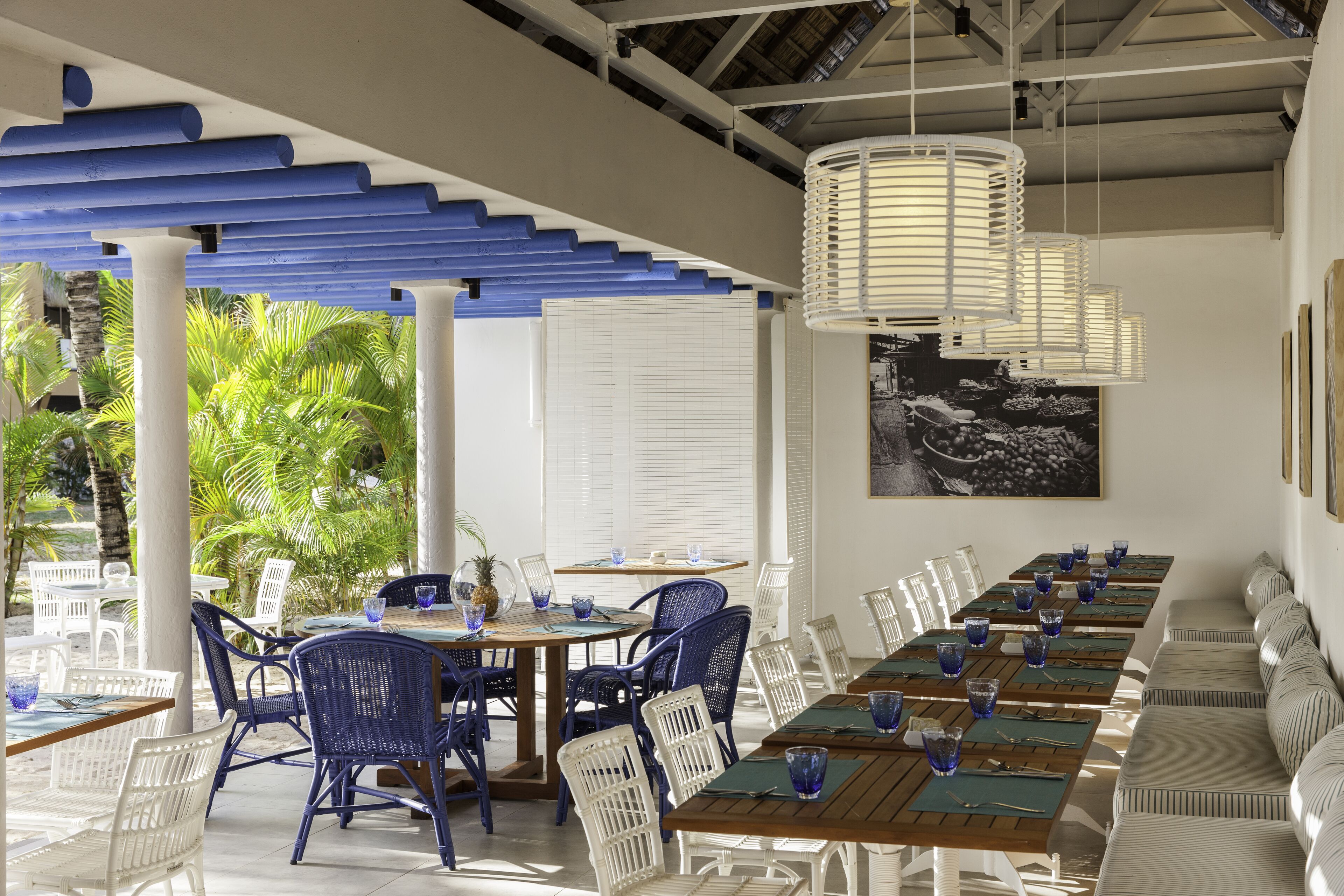 Un espace extérieur abrité, aménagé pour les repas, avec des chaises blanches et bleues, des tables en bois et des luminaires suspendus.