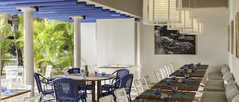 A covered outdoor dining area with white and blue chairs, wooden tables, and hanging light fixtures.