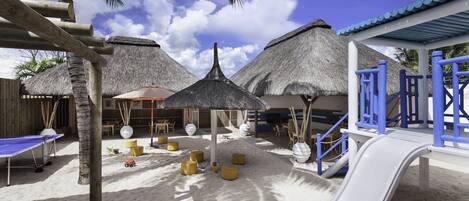 A beachside play area with a slide, thatched-roof structures, and outdoor seating.