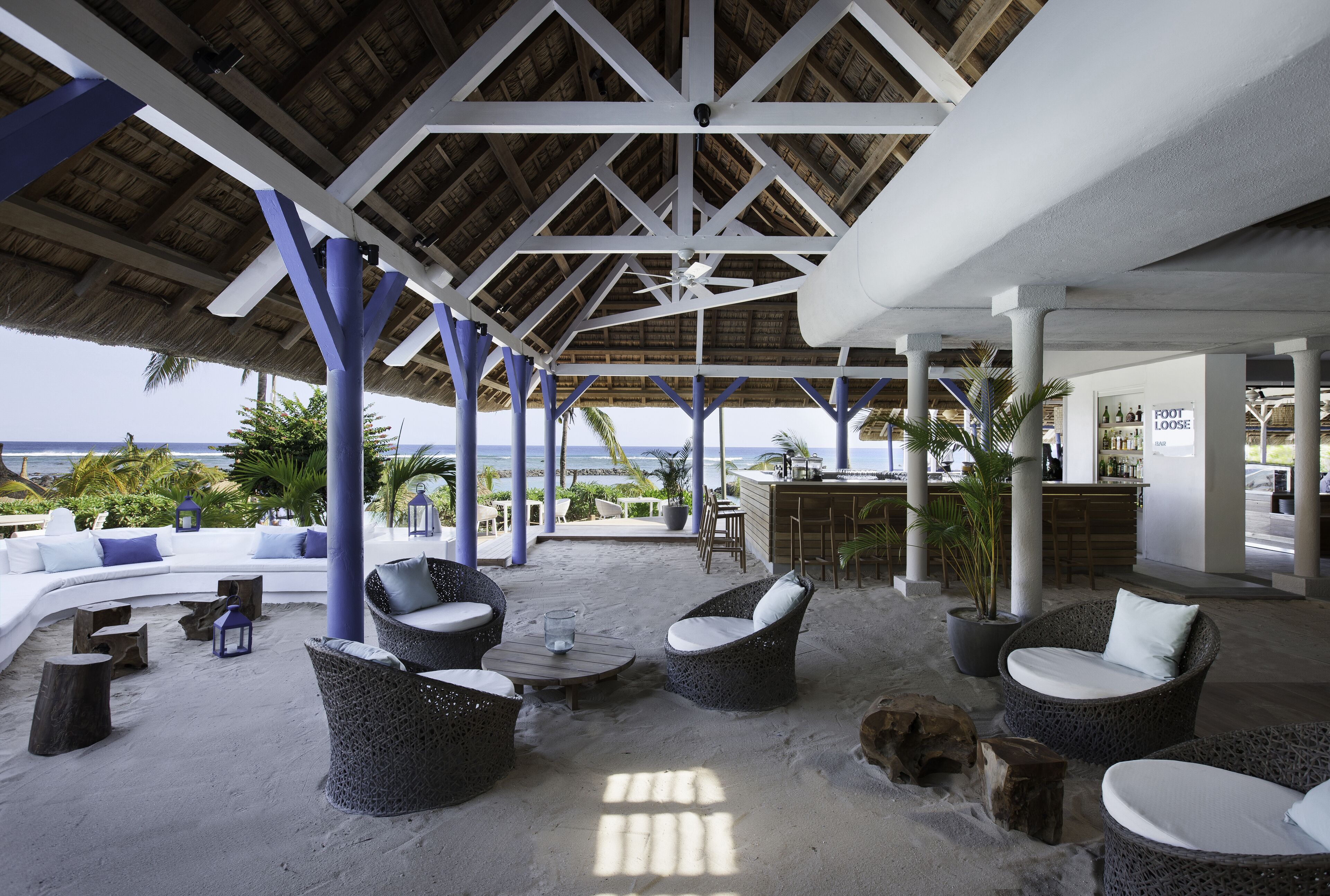 Um lounge à beira-mar com telhado de palha, móveis brancos e azuis, e vista para o oceano.