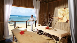 A wooden room with two massage tables, a beach view, and a person in white attire.