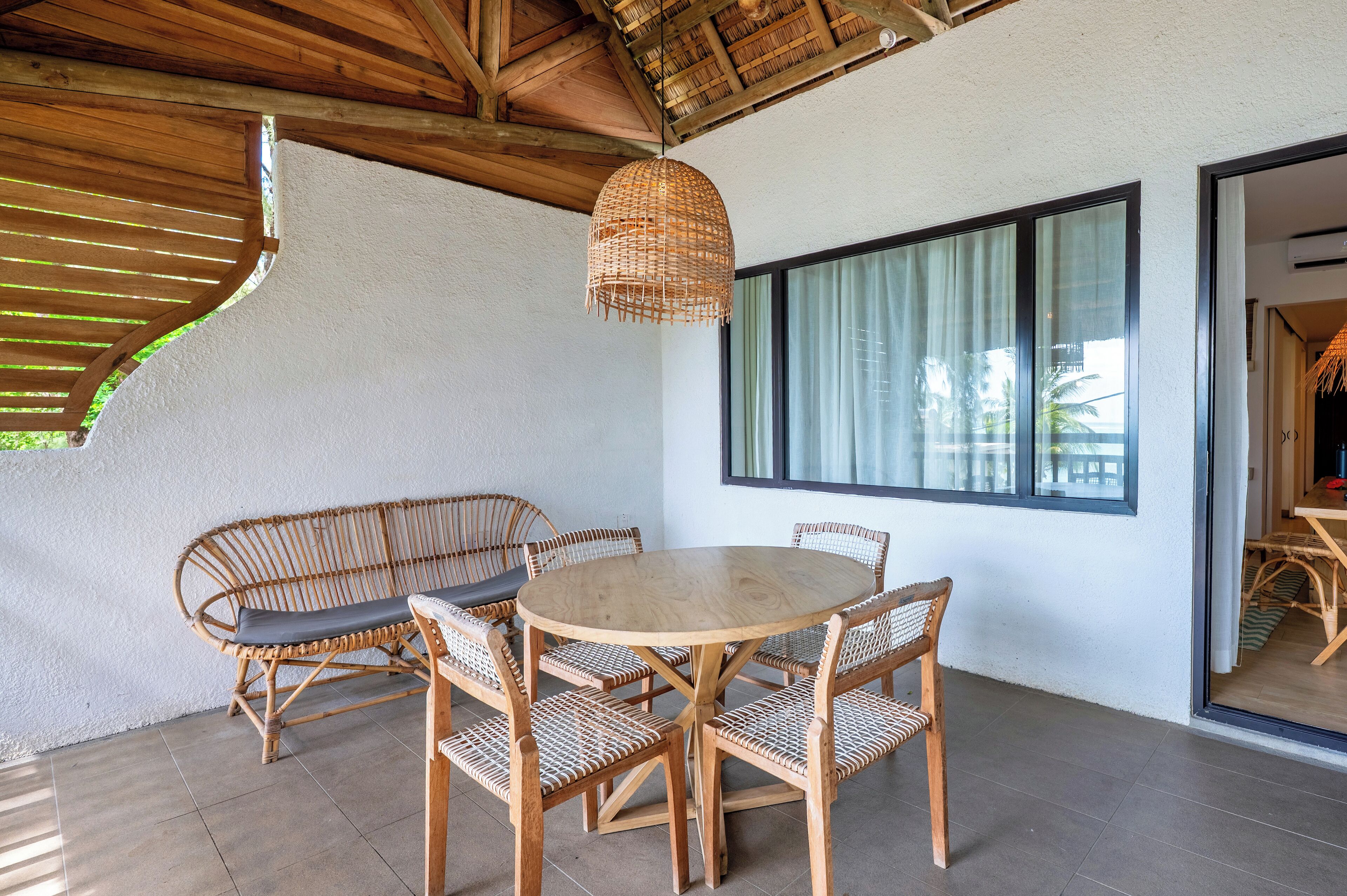 An outdoor patio with a round wooden table surrounded by wicker chairs, a woven pendant light, and a view of the outdoors through large windows.