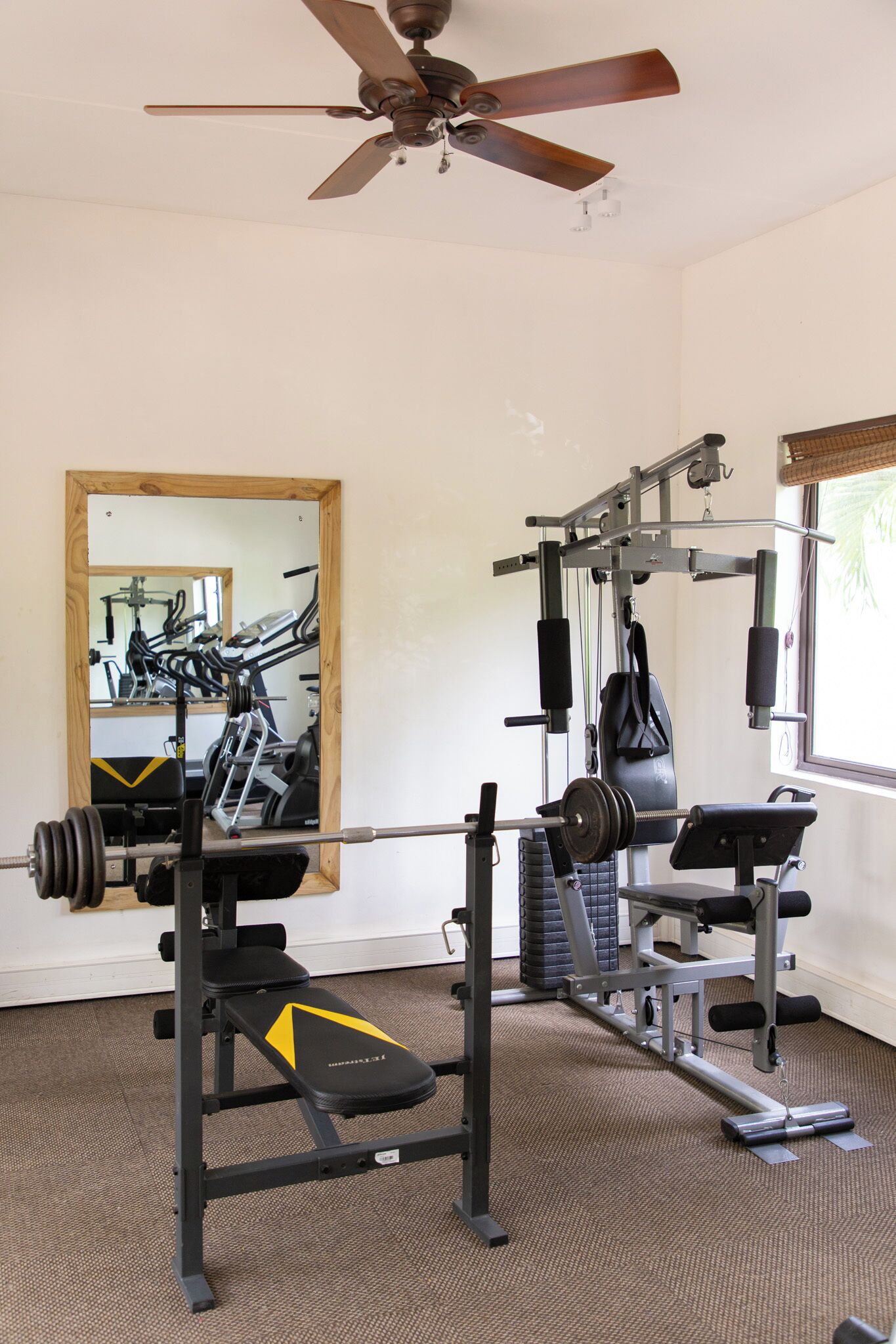 Une salle de sport équipée d’un ventilateur de plafond, d’un miroir et de divers appareils d’exercice, notamment un banc de développé couché et des supports à poids.