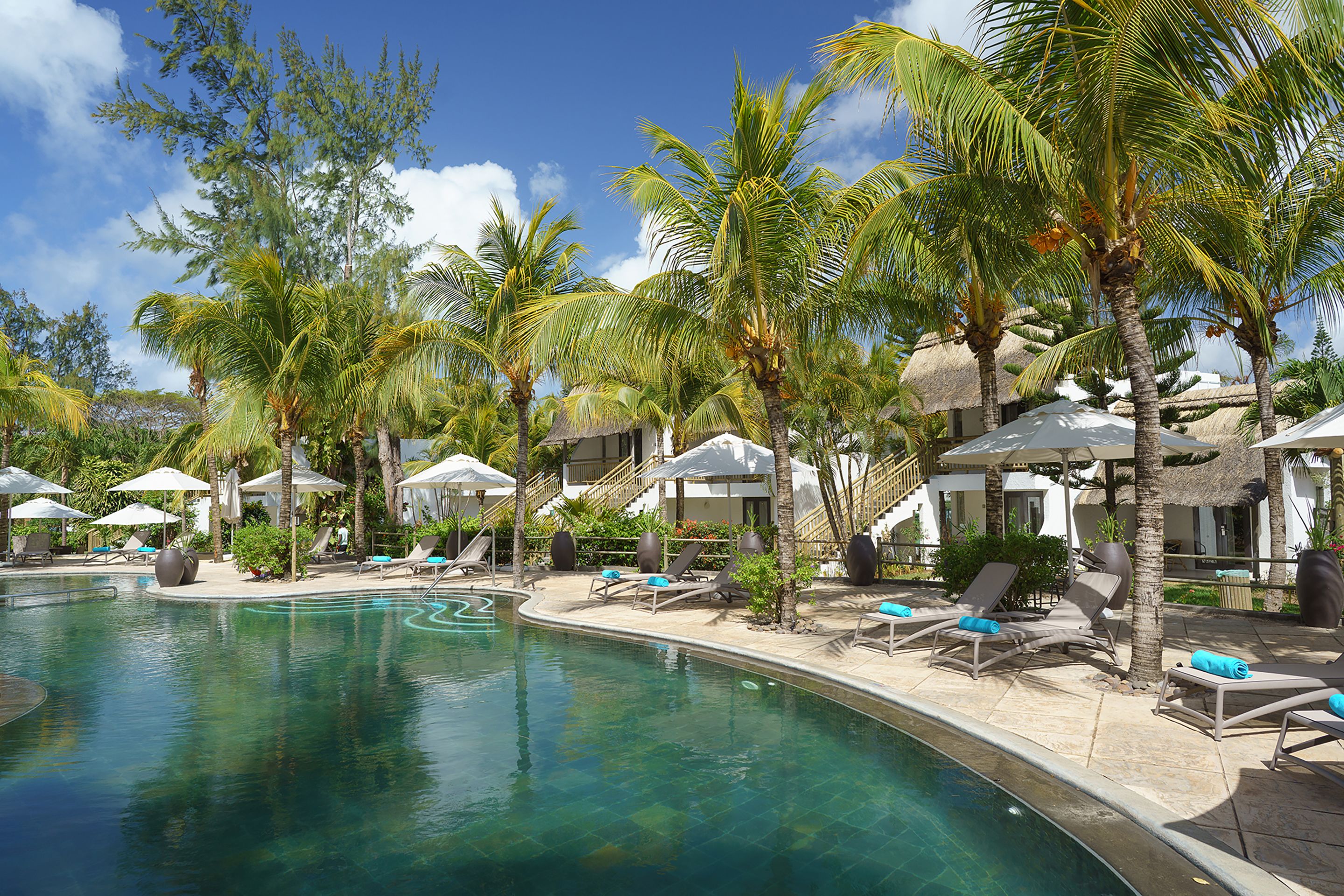 Un área junto a la piscina con sillones de descanso y sombrillas, rodeada de palmeras.