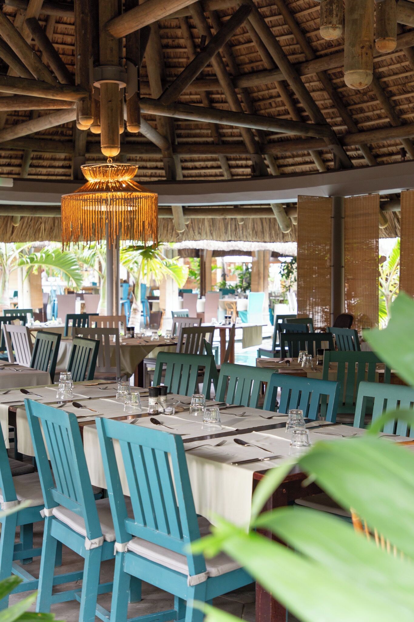 Een restaurant met houten balken en een rieten dak, tafels gedekt met witte tafelkleden en blauwe stoelen, en een hangende kroonluchter.