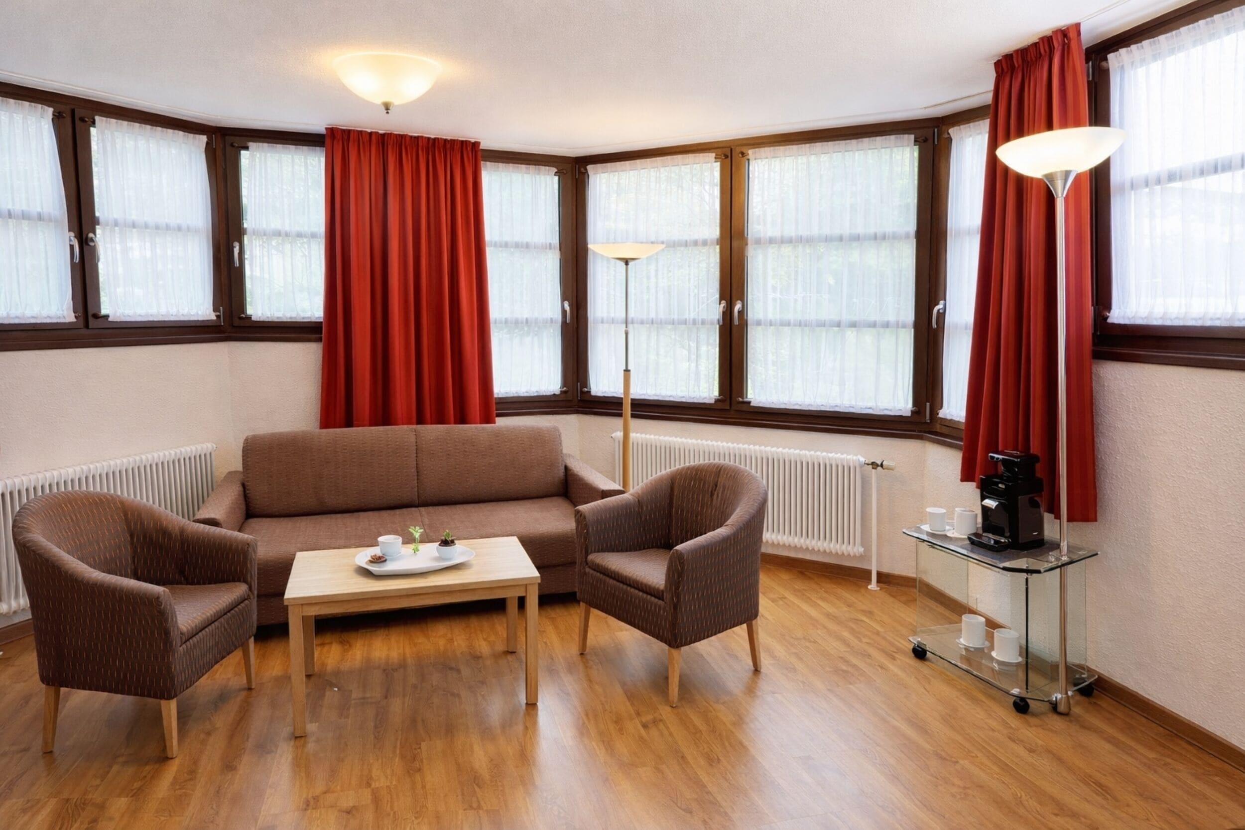 A room with a brown sofa, two armchairs, a small table with a tray, a floor lamp, and a coffee cart.