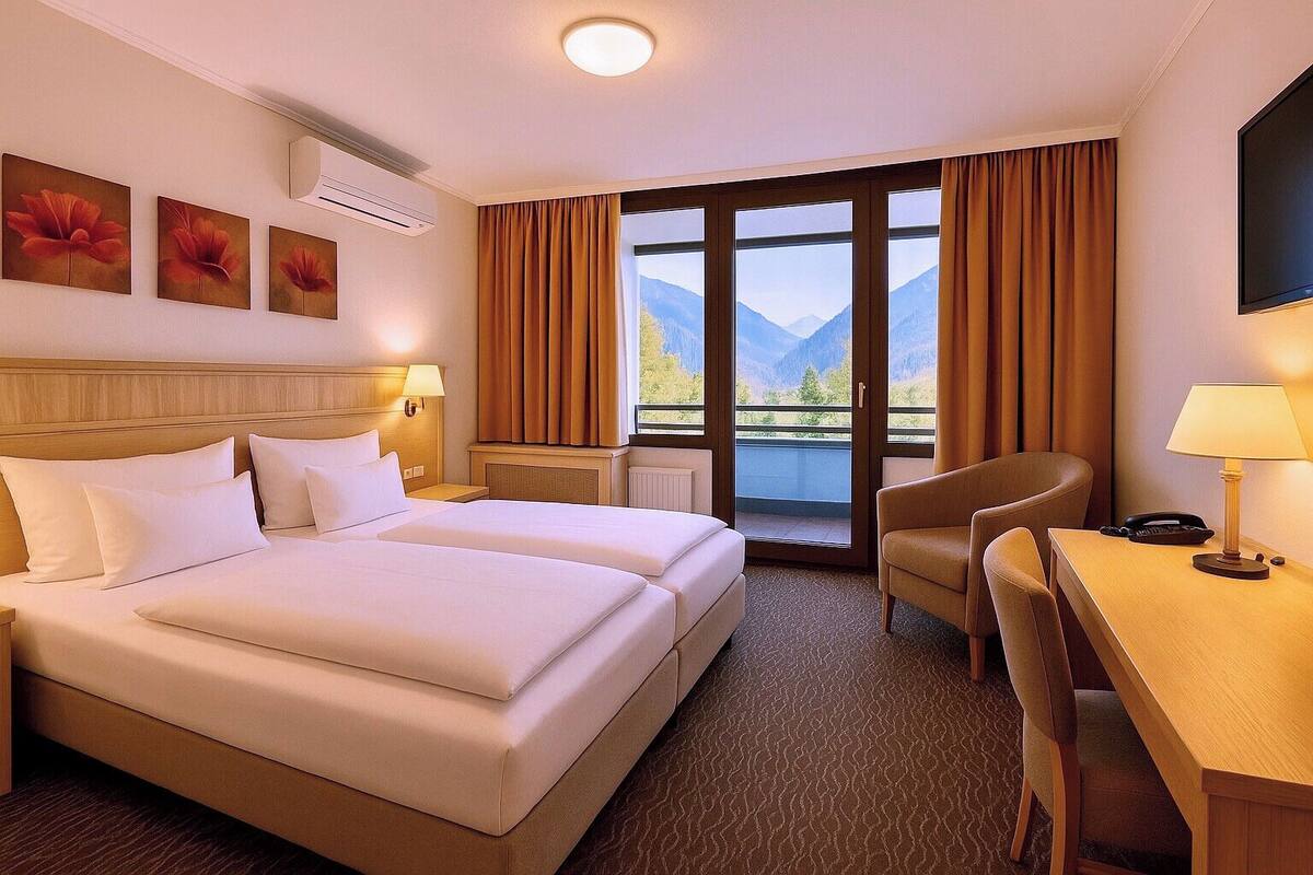 A hotel room with two beds, a desk, a chair, and a large window with a mountain view.