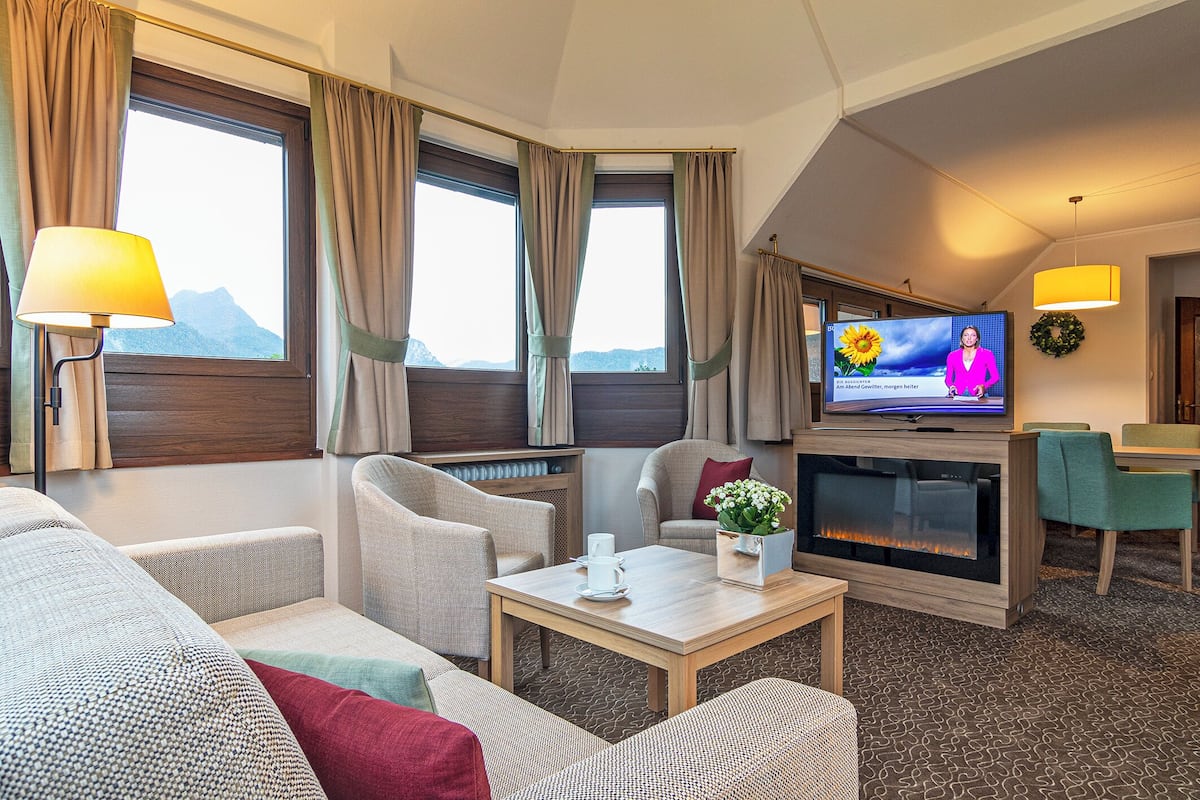 A hotel room with a sofa, armchairs, a television, a fireplace, and a view of mountains through the windows.