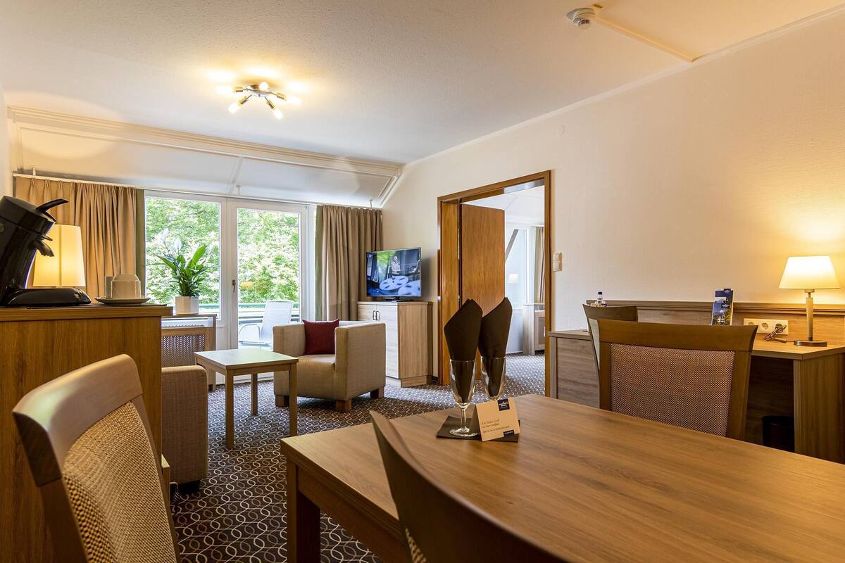 A hotel room with a dining area, a television, and a view of greenery outside.