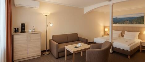 A hotel room with a bed, a sofa, a small table, a desk, a lamp, a cabinet, and a painting on the wall.