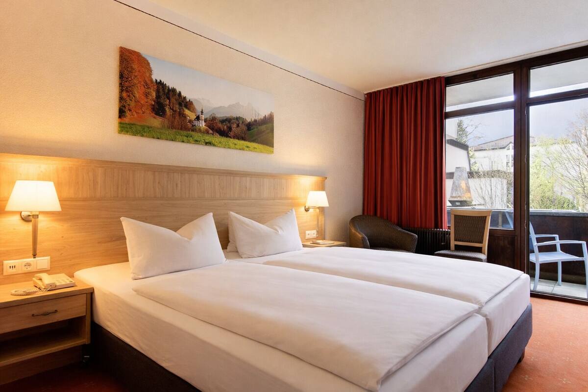 A hotel room with a double bed, two bedside tables with lamps, a chair, a view of the outdoors, and a painting on the wall.