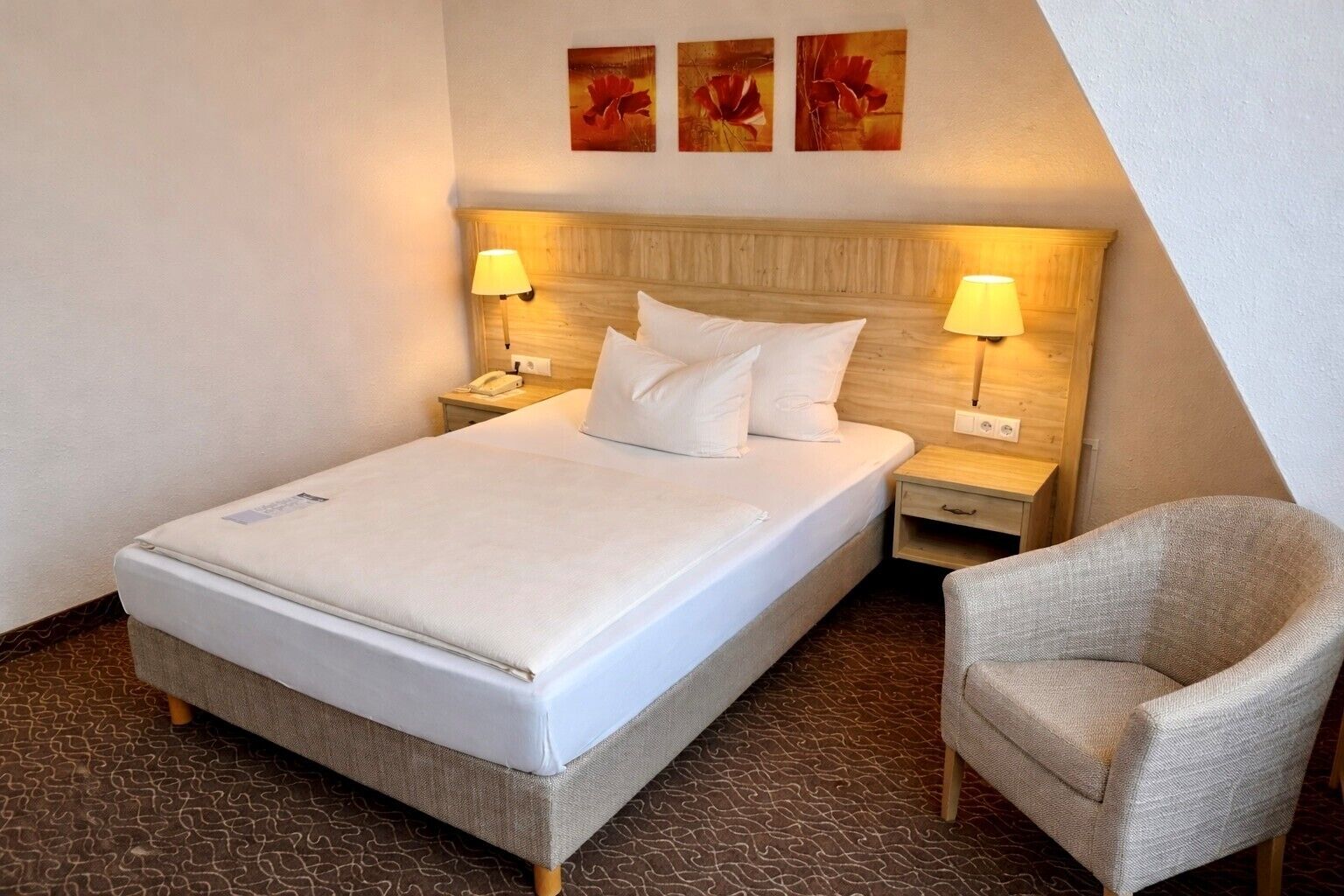 A neatly made bed with white linens, two bedside lamps, a wooden headboard, and a single chair.