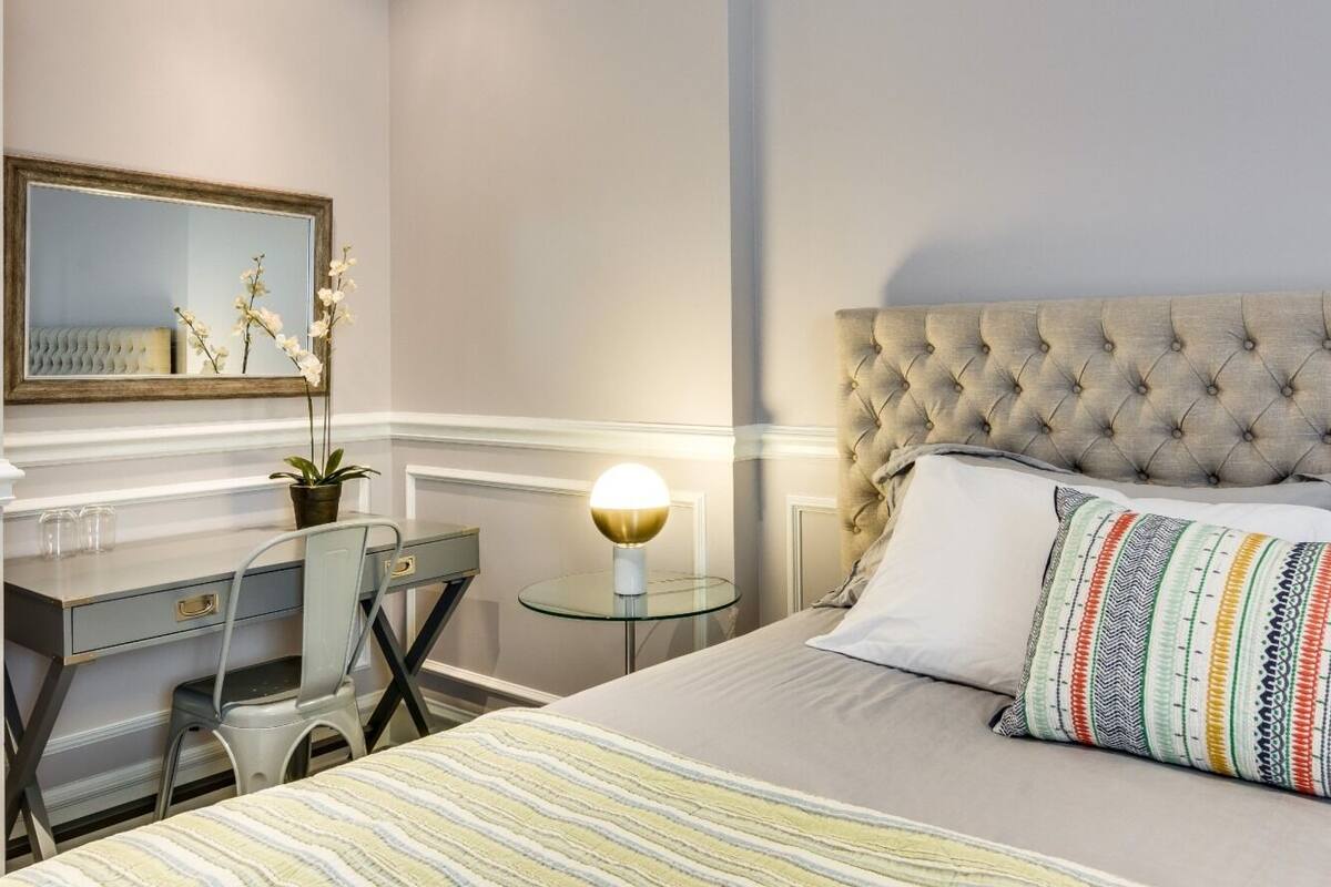 A bedroom with a tufted headboard, a bed with striped bedding, a bedside table with a lamp, a desk with a chair, and a mirror on the wall.