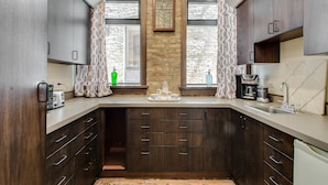 A modern kitchen with dark wood cabinets, a white countertop, and a built-in oven. There are two windows with patterned curtains, a stone accent wall, and a coffee maker on the counter. - The Golden Arm (Chicago)