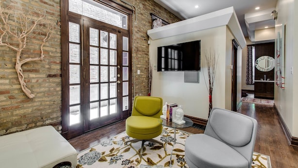 A room with a brick wall, a large wooden door, a flat-screen TV, and a grey armchair. - The Golden Arm (Chicago)