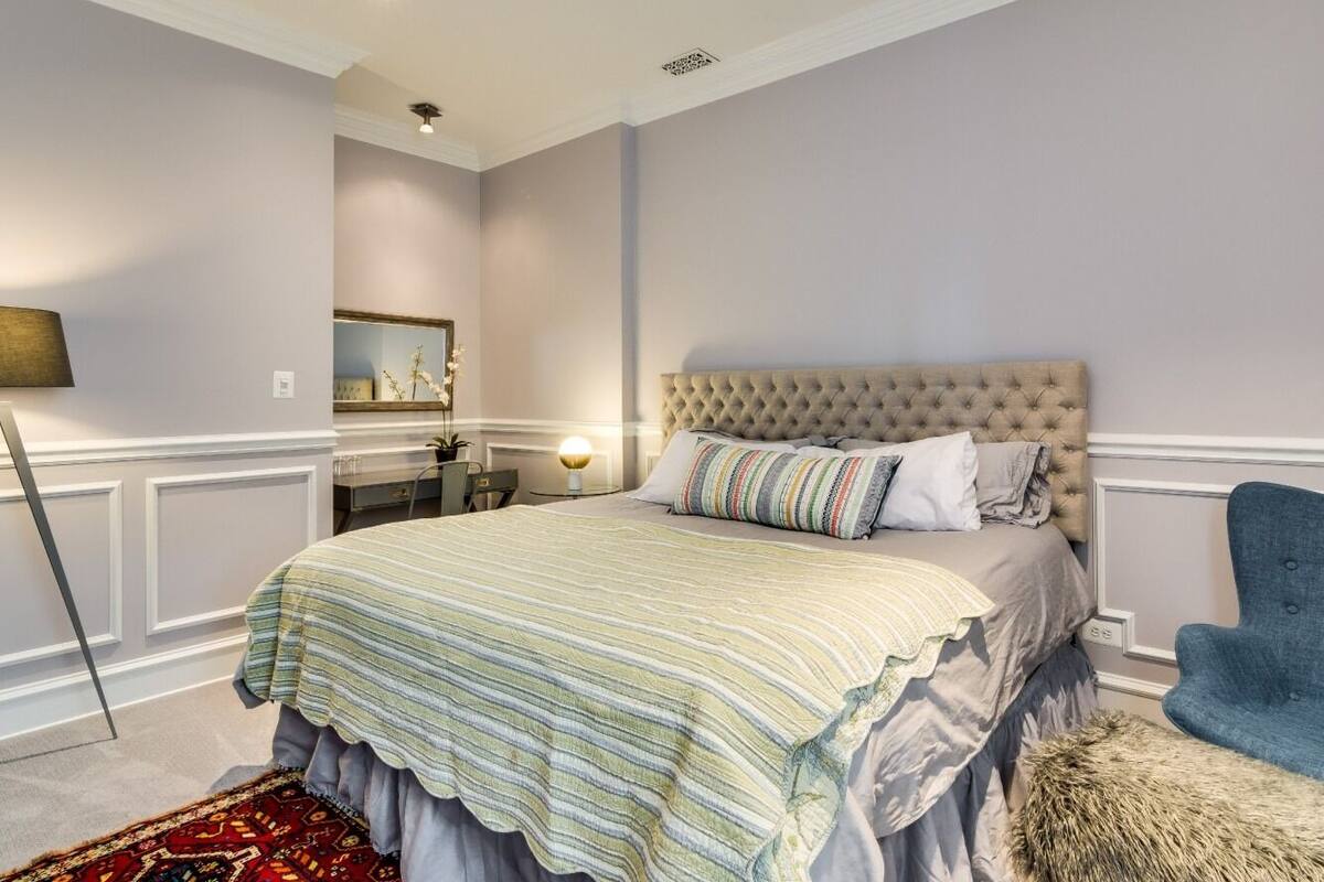 A bedroom with a tufted headboard, a bed with striped bedding, a blue armchair, and a small side table with a lamp.