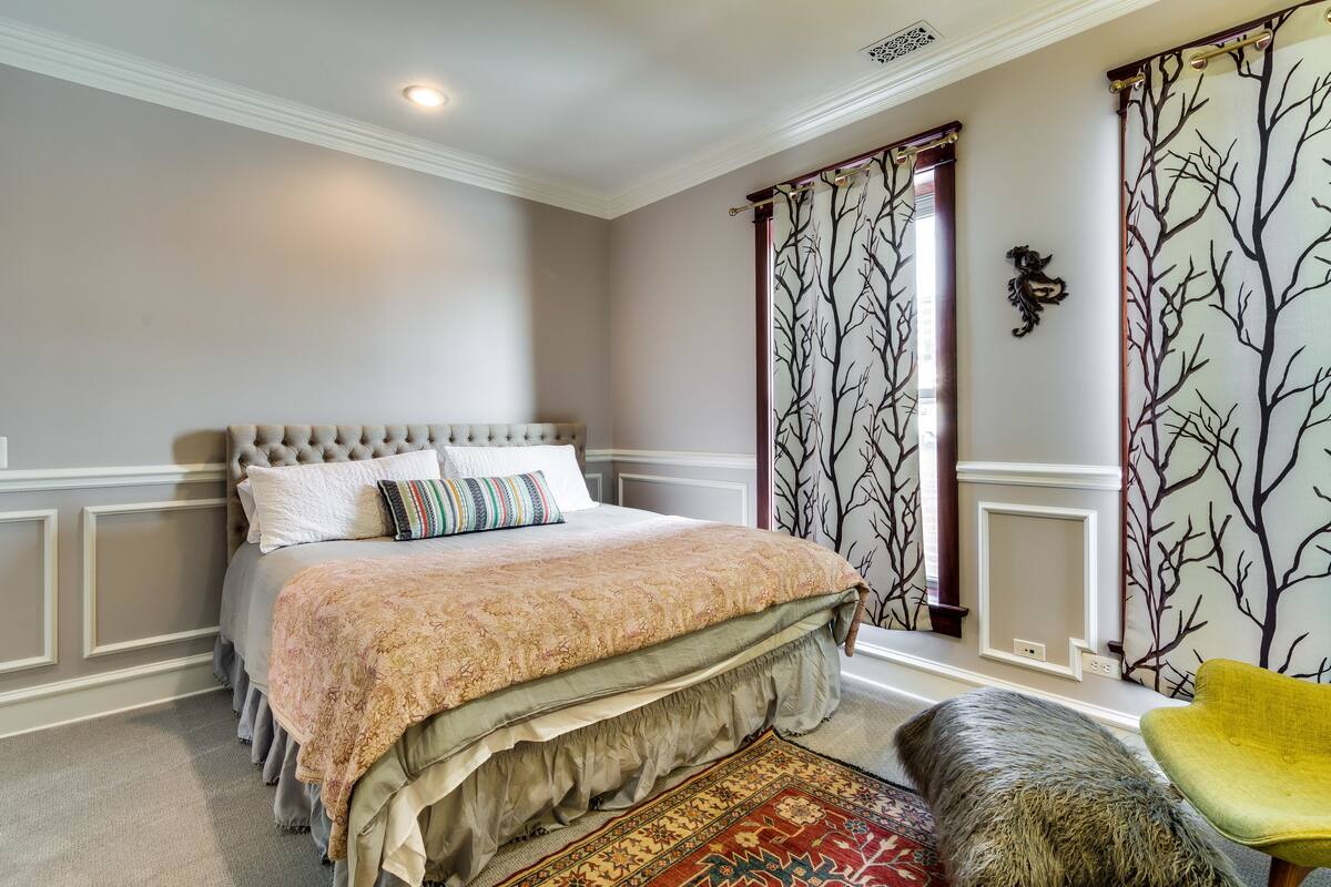 A bedroom with a tufted headboard, a large bed with a patterned bedspread, a decorative throw, a ruffled skirt on the bed, a red and yellow patterned rug, a yellow chair, and a tree-patterned curtain.