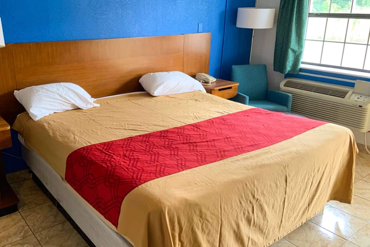 A hotel room with a wooden headboard, a bed with a beige and red bedspread, two bedside lamps, a nightstand, a blue chair, and a window with curtains.