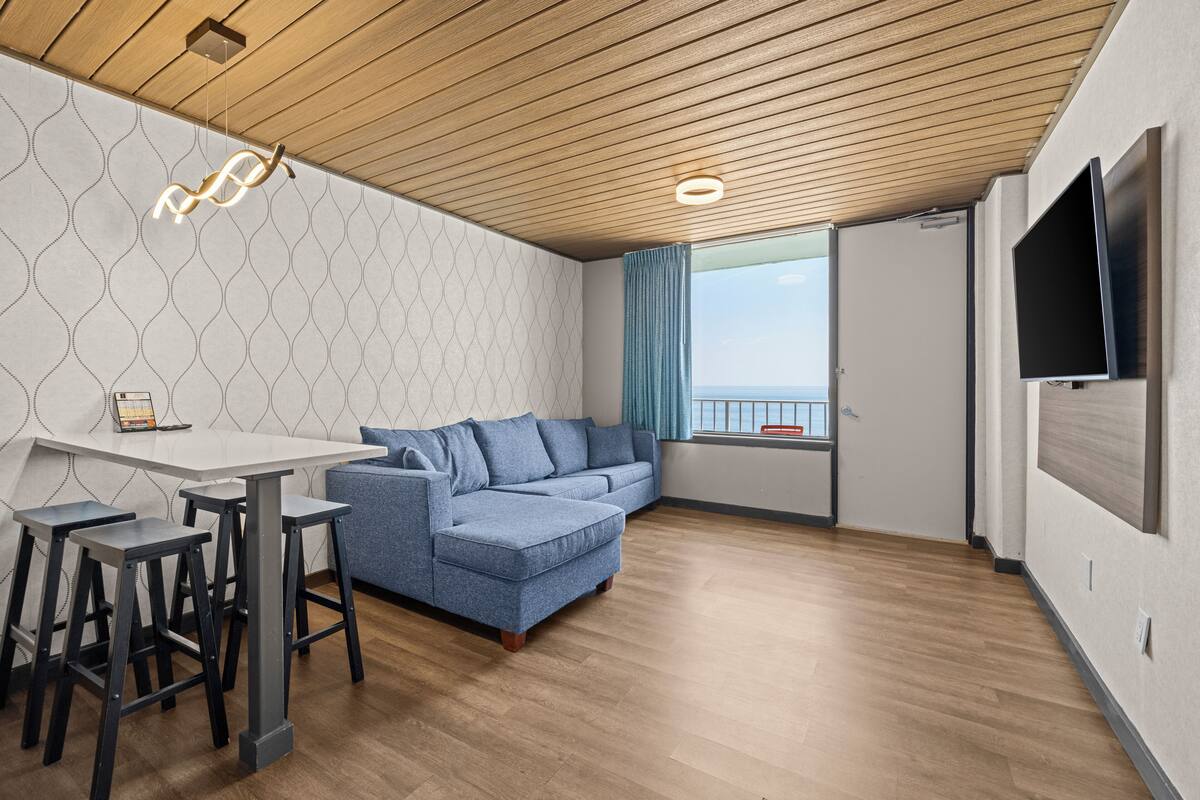 A modern living room with a wooden ceiling, a blue sofa, a white table with bar stools, a TV, and a window with a view.