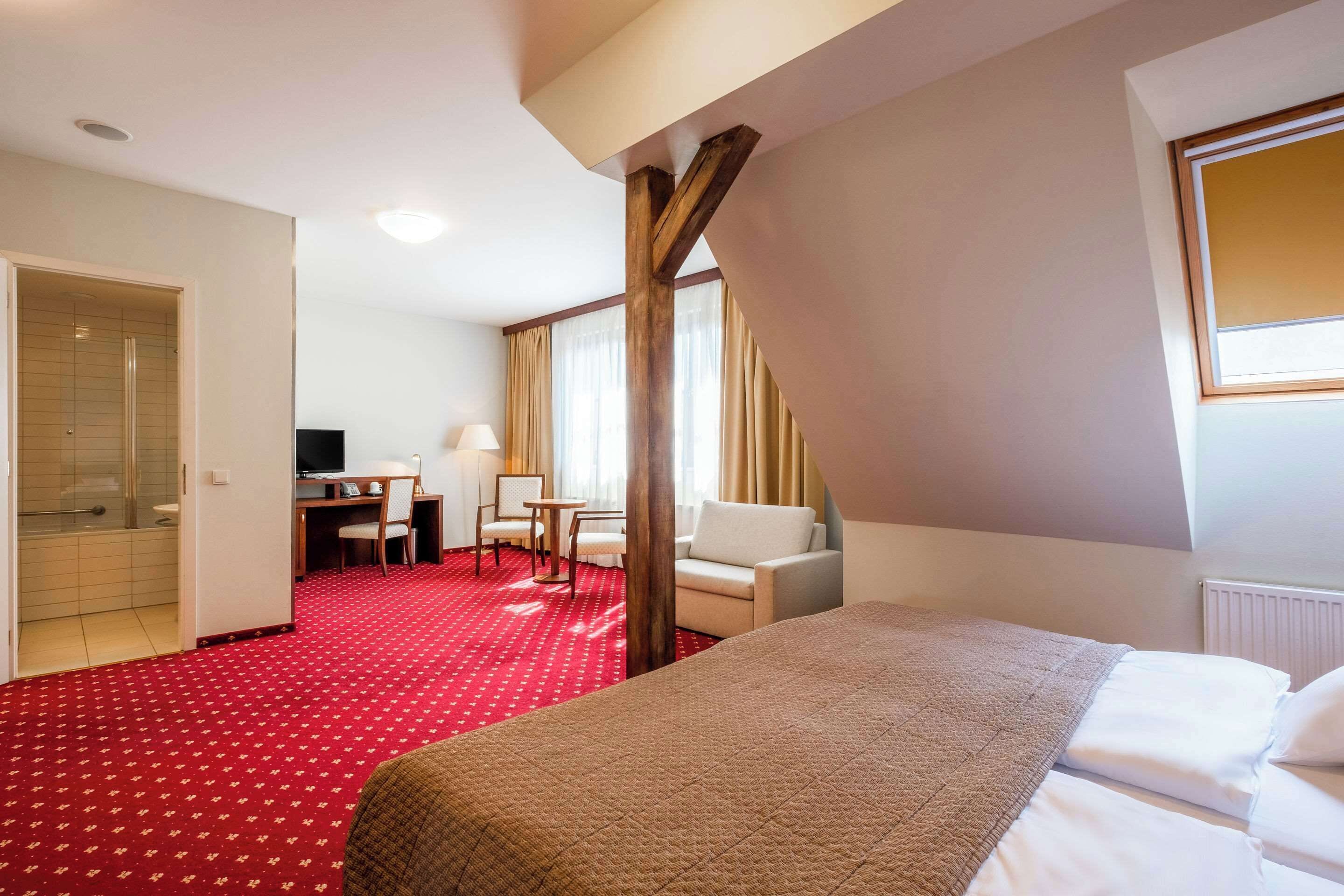 A hotel room with a red carpet, a bed, a desk with a chair, a sofa, and a window with curtains.