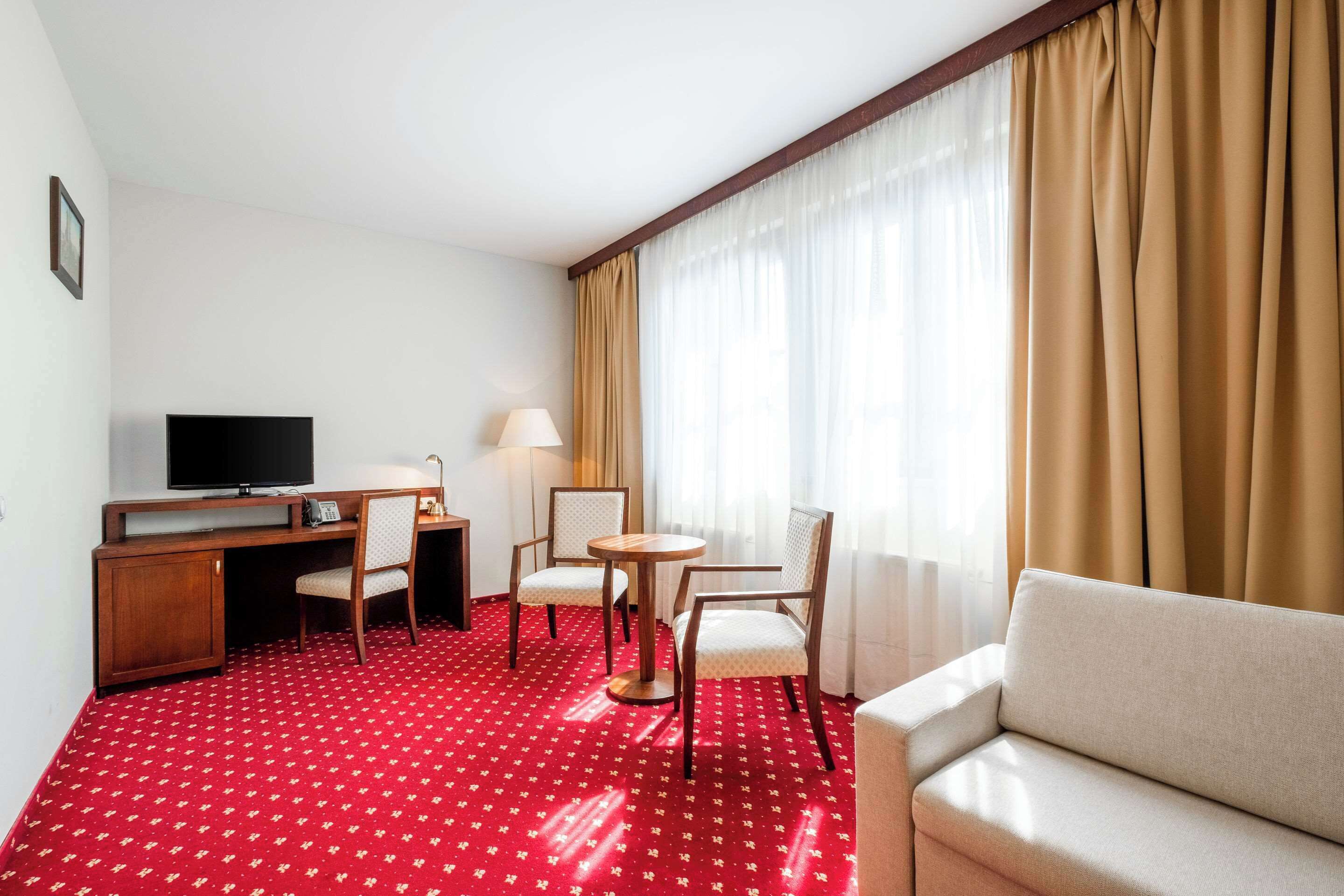 A hotel room with a red carpet, a sofa, a desk with a television, two chairs, a lamp, and a large window with curtains.