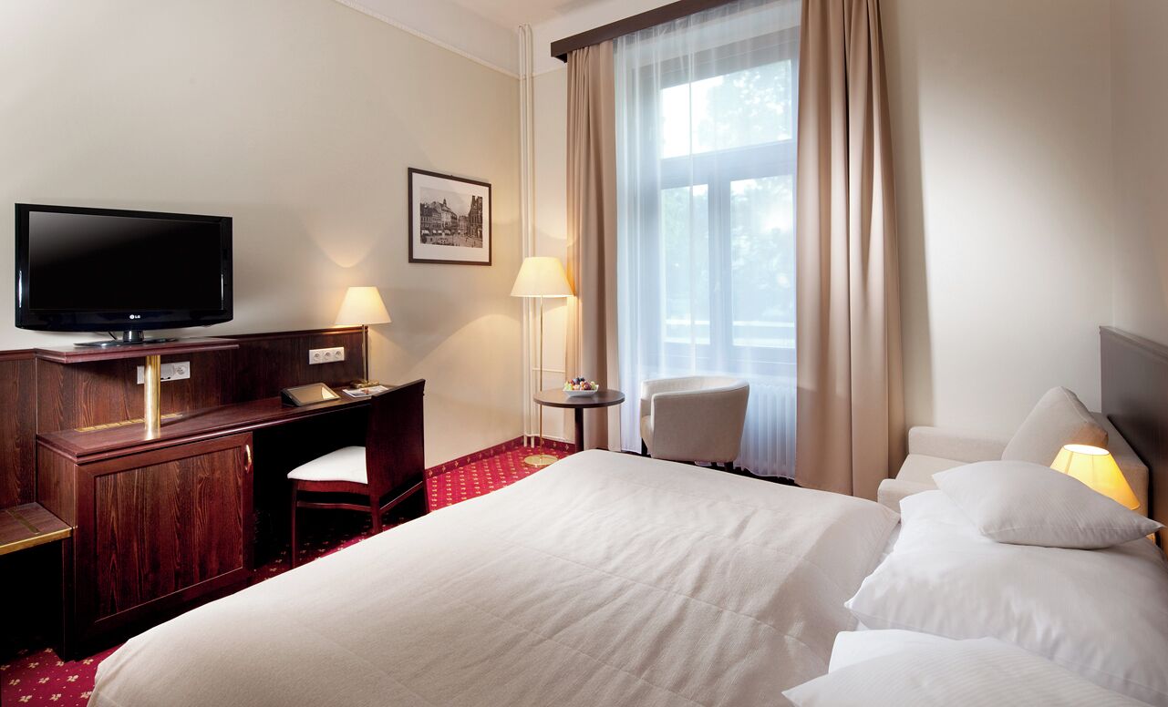 A hotel room with a large bed, a desk with a chair, a television, a lamp, and a window with curtains.