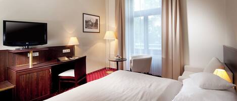 A hotel room with a large bed, a desk with a chair, a television, a lamp, and a window with curtains.