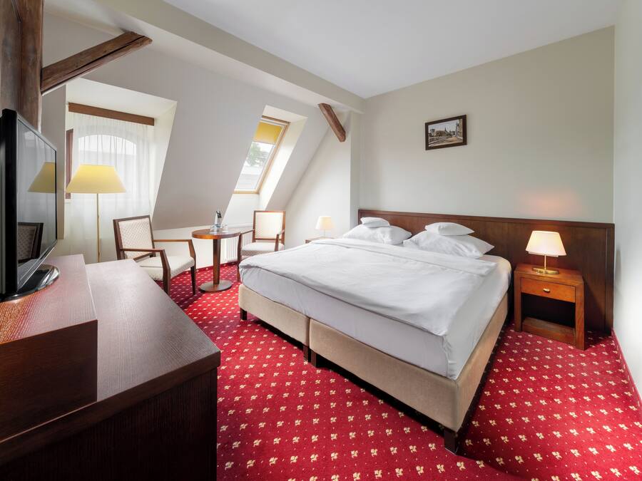 A hotel room with a large bed, a desk, a chair, a small table, and a lamp.