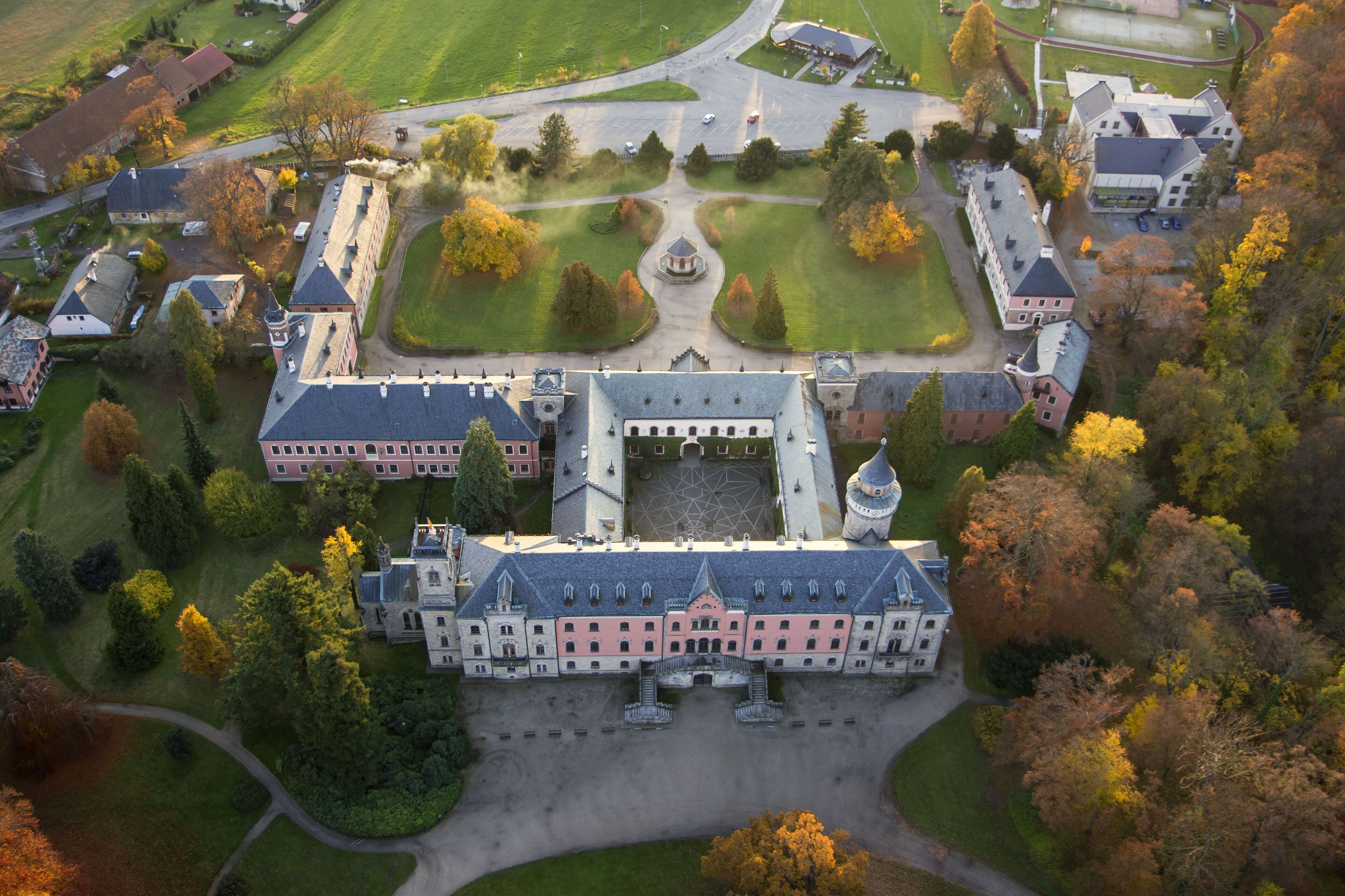 Une vue aérienne d’un grand bâtiment historique, doté de plusieurs ailes, entouré de jardins et d’arbres bien entretenus.