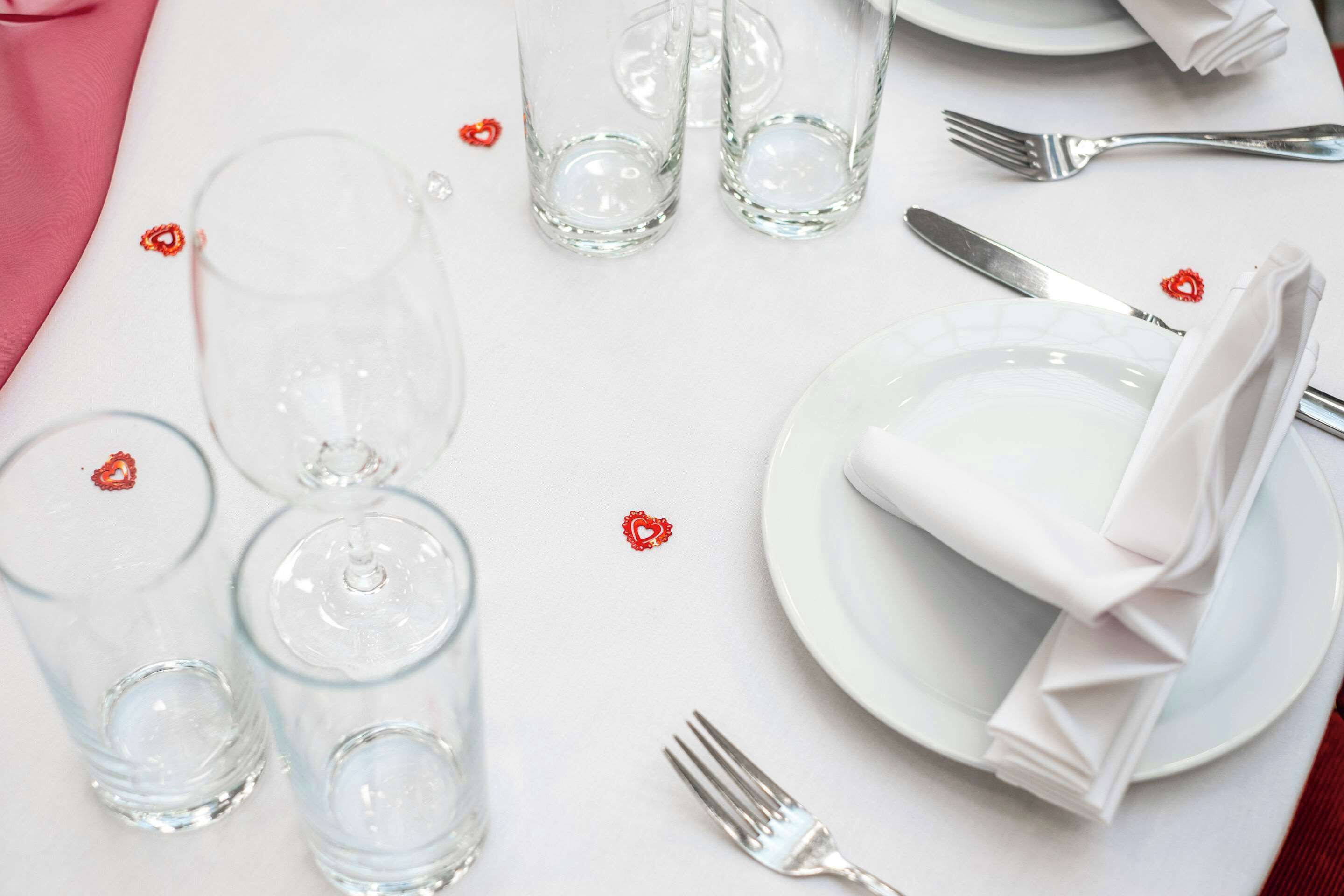 Una mesa dispuesta con vasos traslúcidos, platos y servilletas dobladas, con pequeñas decoraciones en forma de corazón rojo.