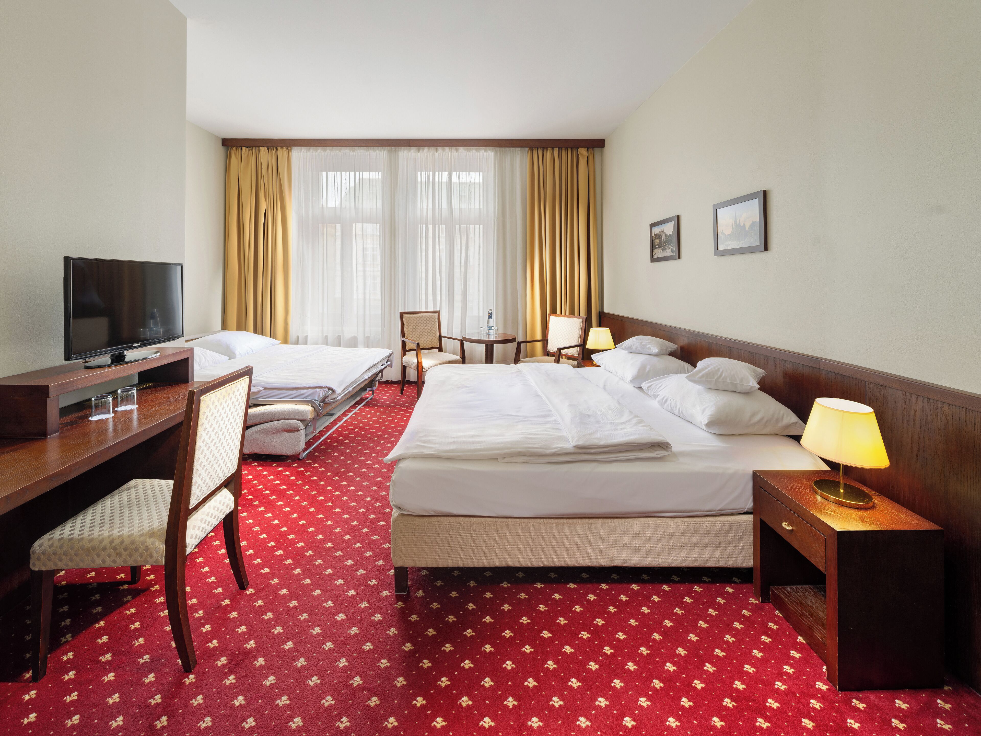 Habitación de hotel con dos camas, un escritorio con televisor, una silla, una mesita de noche con lámpara y alfombra roja.