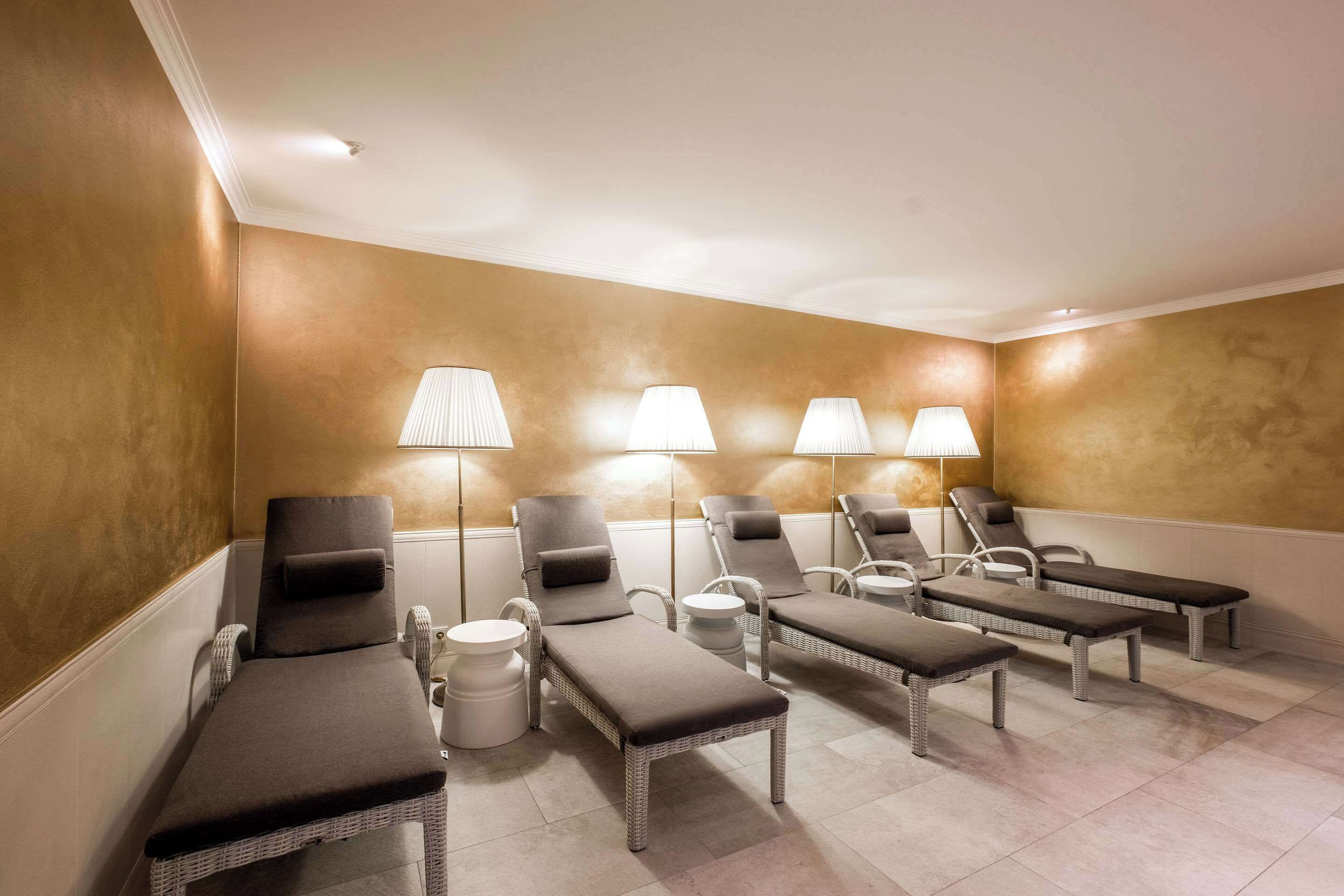 Un salón de spa con seis sillones reclinables, cada uno con una almohada y una mesita. Tres lámparas fijadas a la pared brindan iluminación. El ambiente tiene suelo de baldosas color beige y paredes con textura.