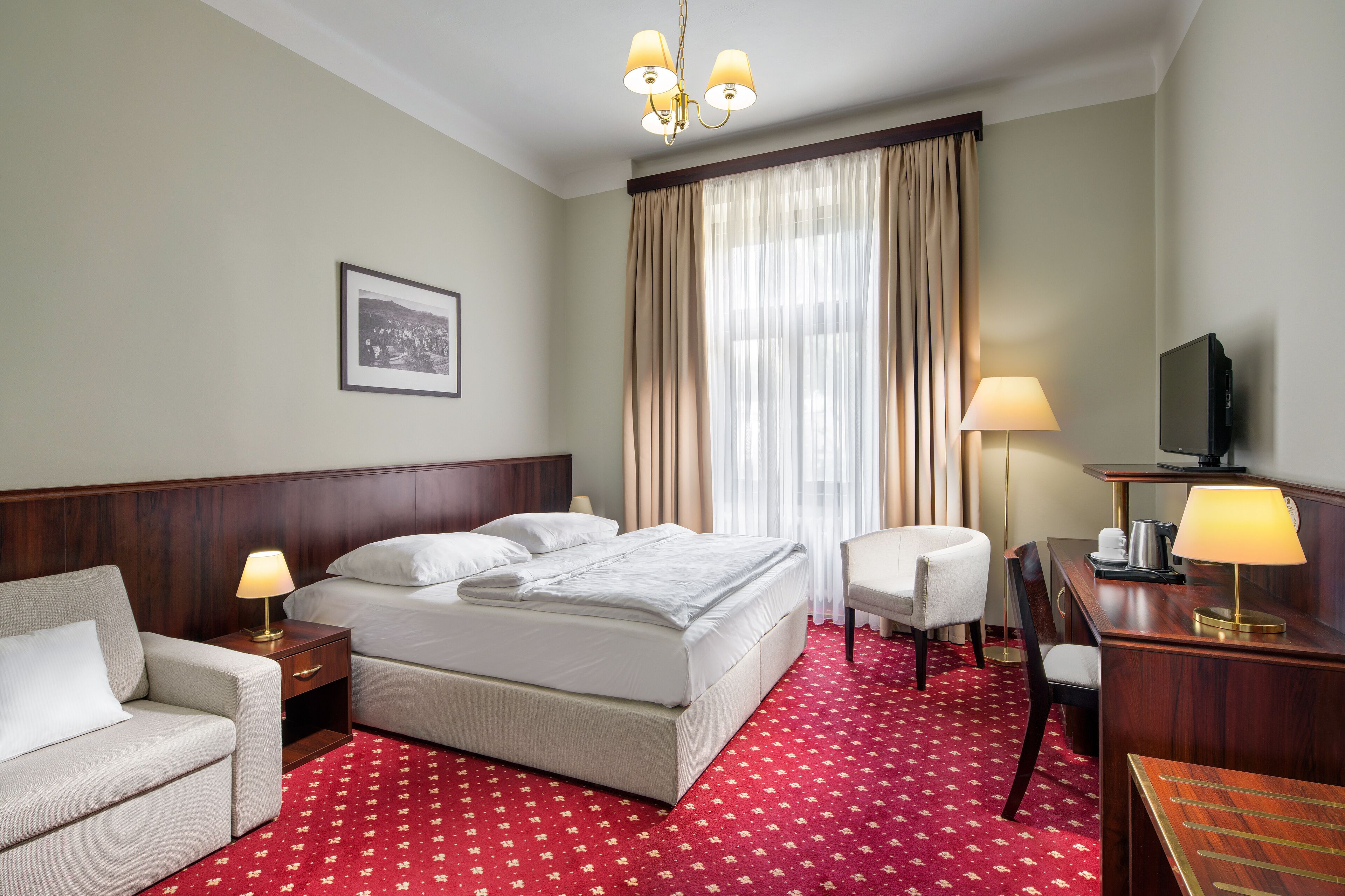 Una habitación de hotel con cama, sofá, escritorio con lámpara, televisión y un ventanal con cortinas.