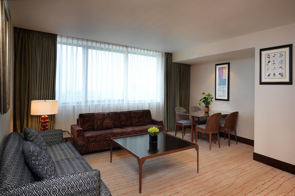 Habitación de hotel con sofá, mesa de centro, mesa de comedor con sillas, lámpara y ventana con cortinas.