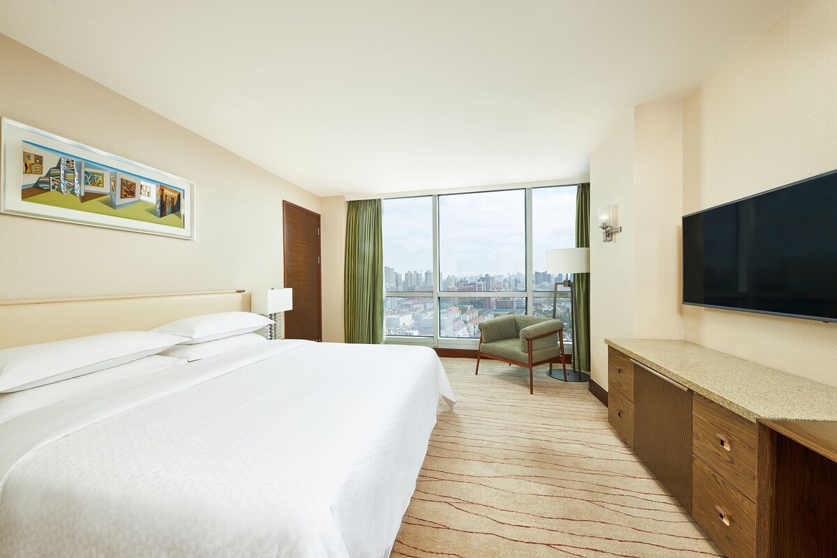 Habitación de hotel con una cama grande, un escritorio, un televisor de pantalla plana, una silla y un ventanal con vista a la ciudad.